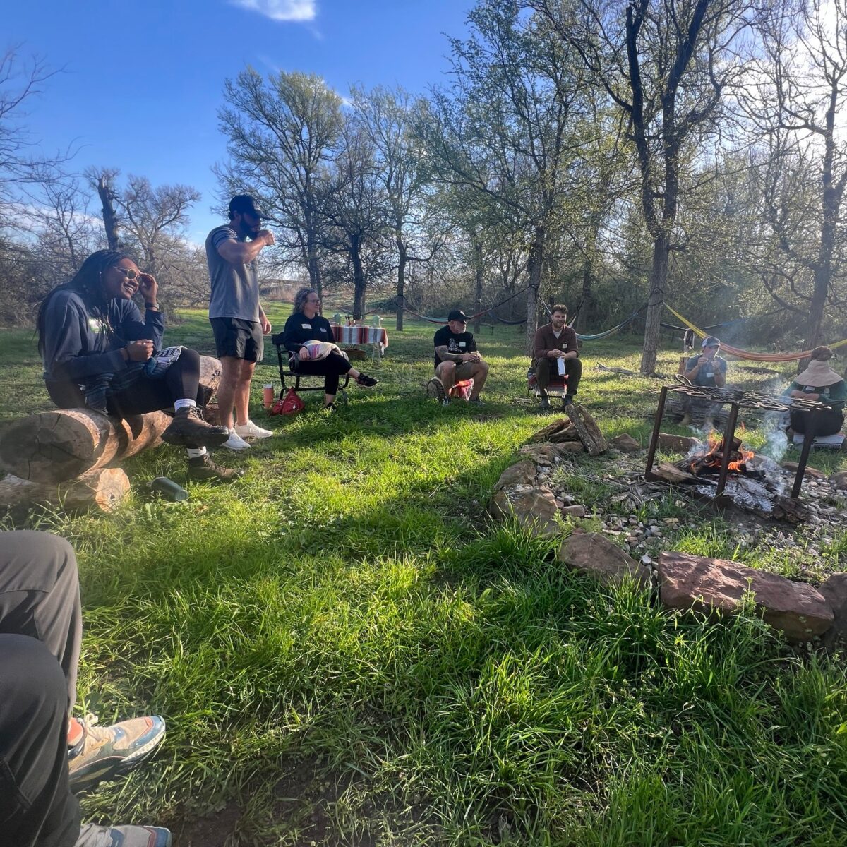 A group of people gathered in a sunny clearing with some sitting and others standing near a fire pit