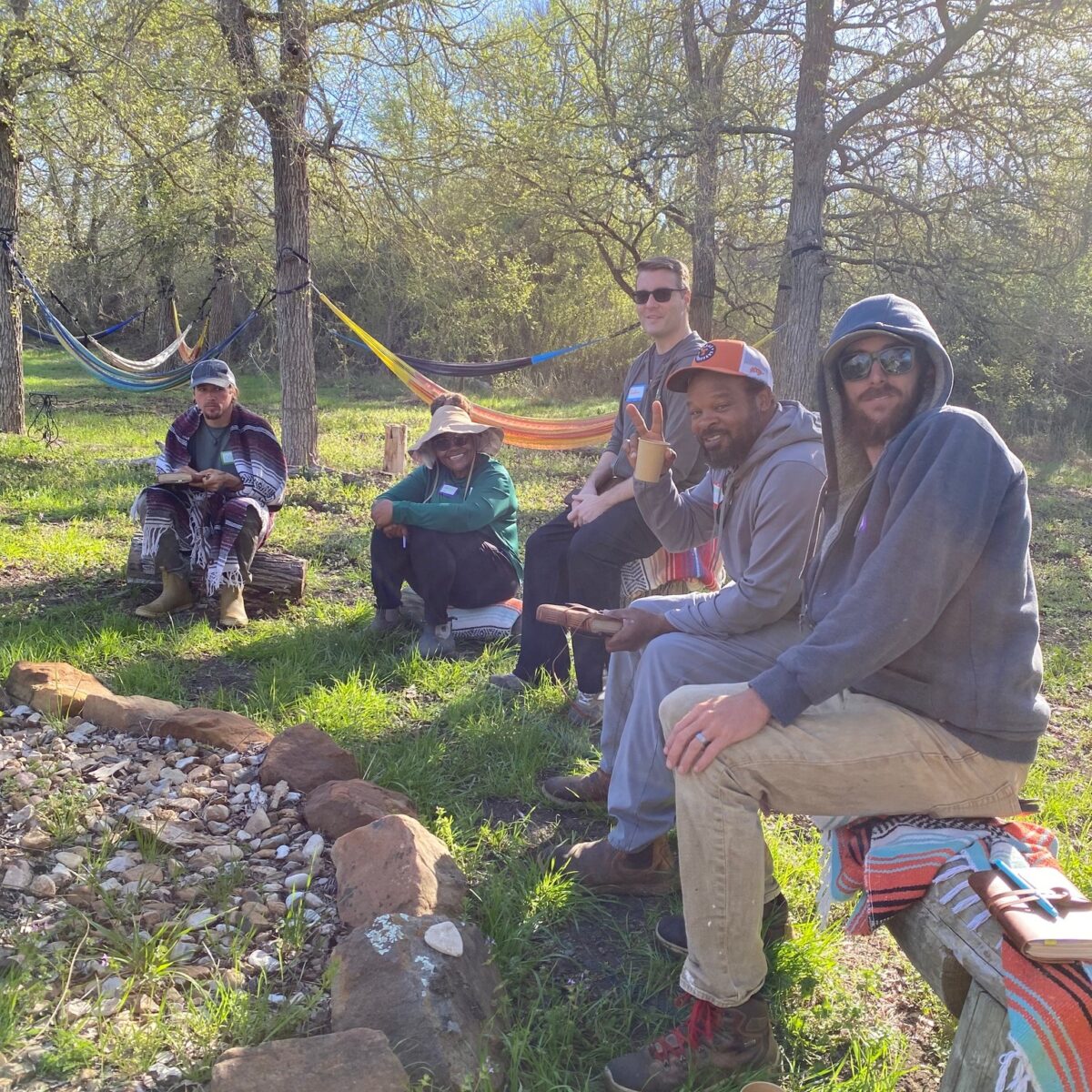 Several people sitting outdoors around a fire pit surrounded by trees and greenery.