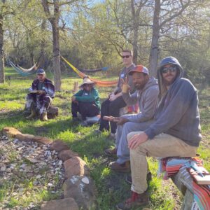 Several people sitting outdoors around a fire pit surrounded by trees and greenery.