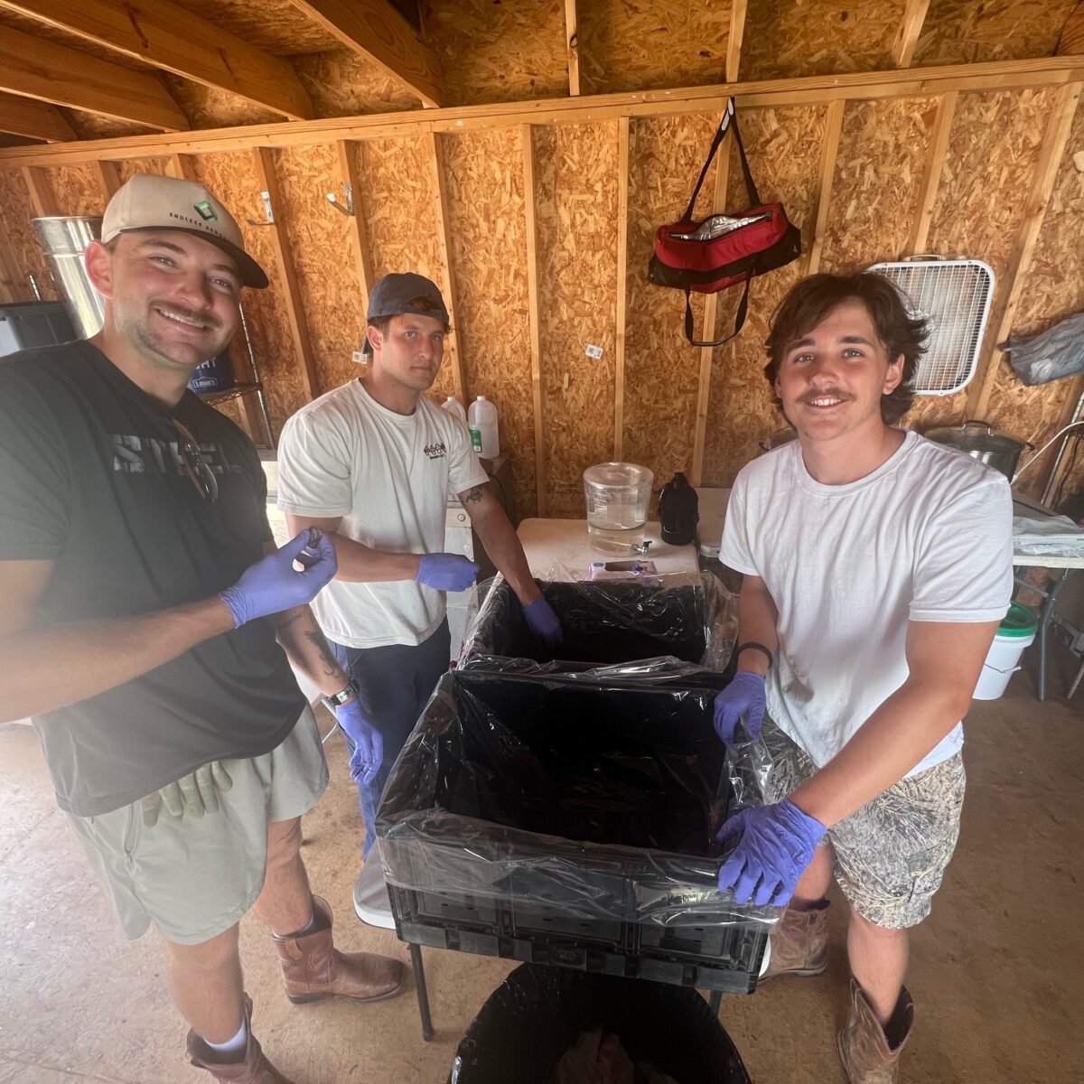 Three people standing inside a rustic wooden shed with a large black cast iron pot on a tripod in front of them.
