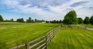 A scenic view of a green pasture with a wooden fence running diagonally under a partly cloudy sky.